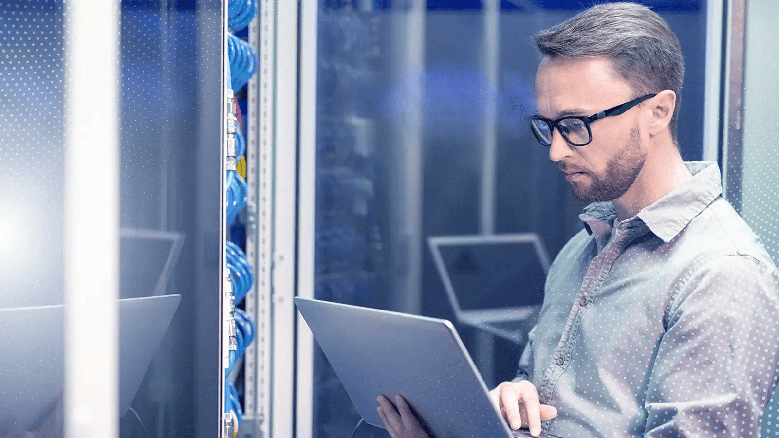 IT professional working on a laptop in a server room with networking cables and equipment racks in the background.