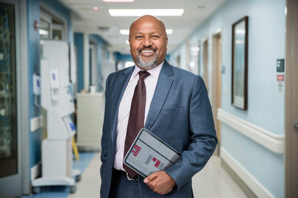 A professional man in a suit stands in a hospital corridor, holding a tablet displaying data, surrounded by medical facilities.