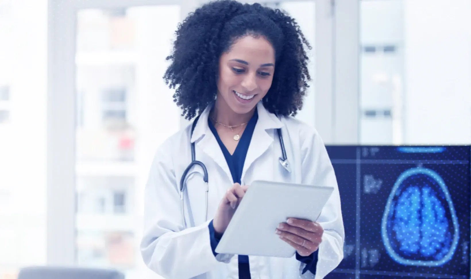 A doctor in a white coat with a stethoscope holds a tablet, standing in a bright room with a brain scan image on a screen.