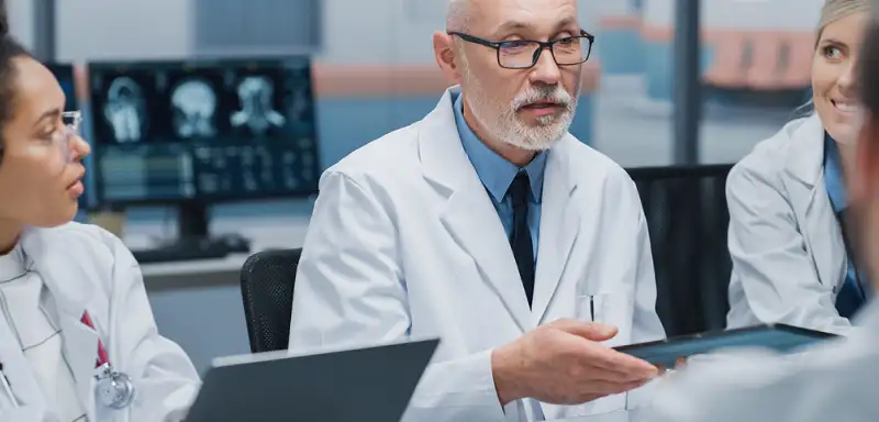 A group of doctors in white coats convenes at a conference table, discussing medical data displayed on a laptop and screens.
